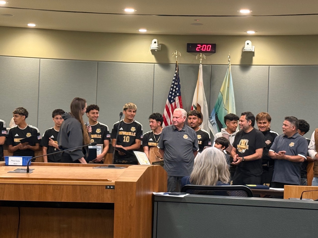 Watsonville High boys' soccer team and their coaches stand in a formal city council chamber while receiving certificates of recognition during an awards ceremony.