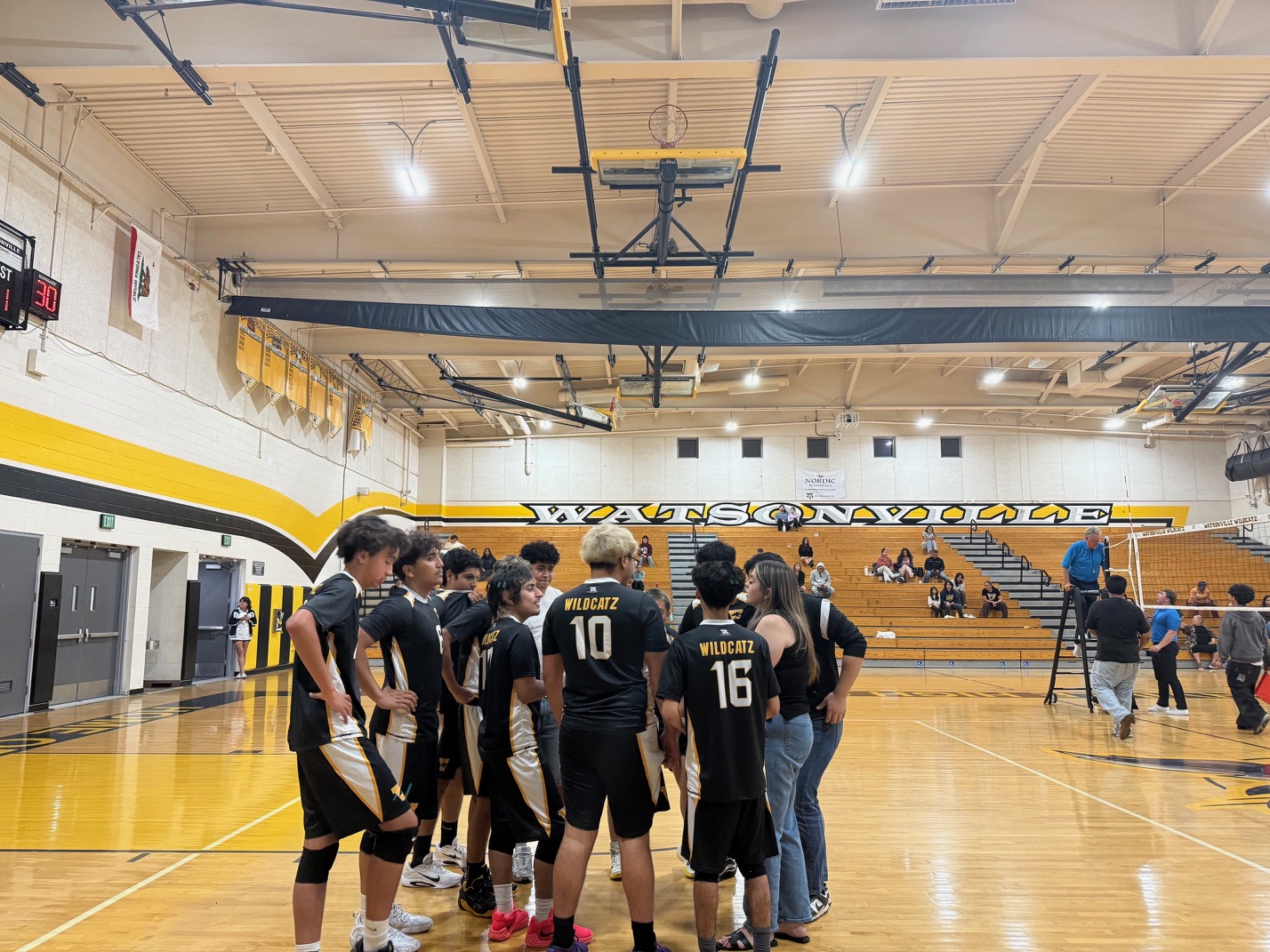 Watsonville High boys' volleyball team in black and gold jerseys huddles together on an indoor basketball court during a match.